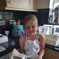 Girl with apron in kitchen holding decorated cupcakes and birthday cake in background