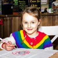 Girl wearing rainbow coloured cardigan sat in cafe colouring a teatowel