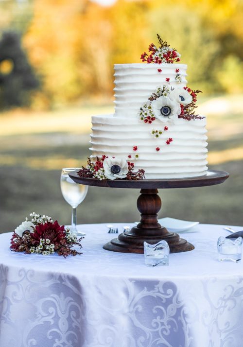 Two tier white buttercream wedding cake with ribbed pattern, white and red flowers on a table outside
